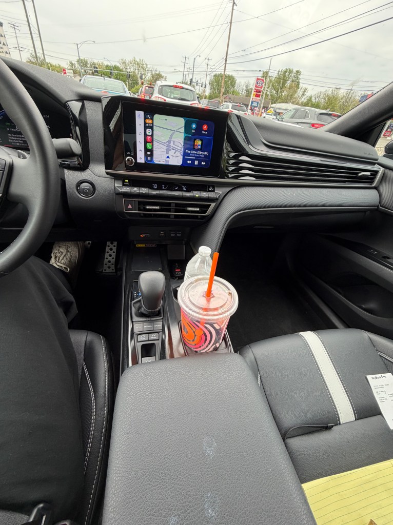 View of a car interior showing the dashboard with a touchscreen display, gear shifter, and a drink in a cup holder. The vehicle is in a traffic situation with other cars visible in the background.