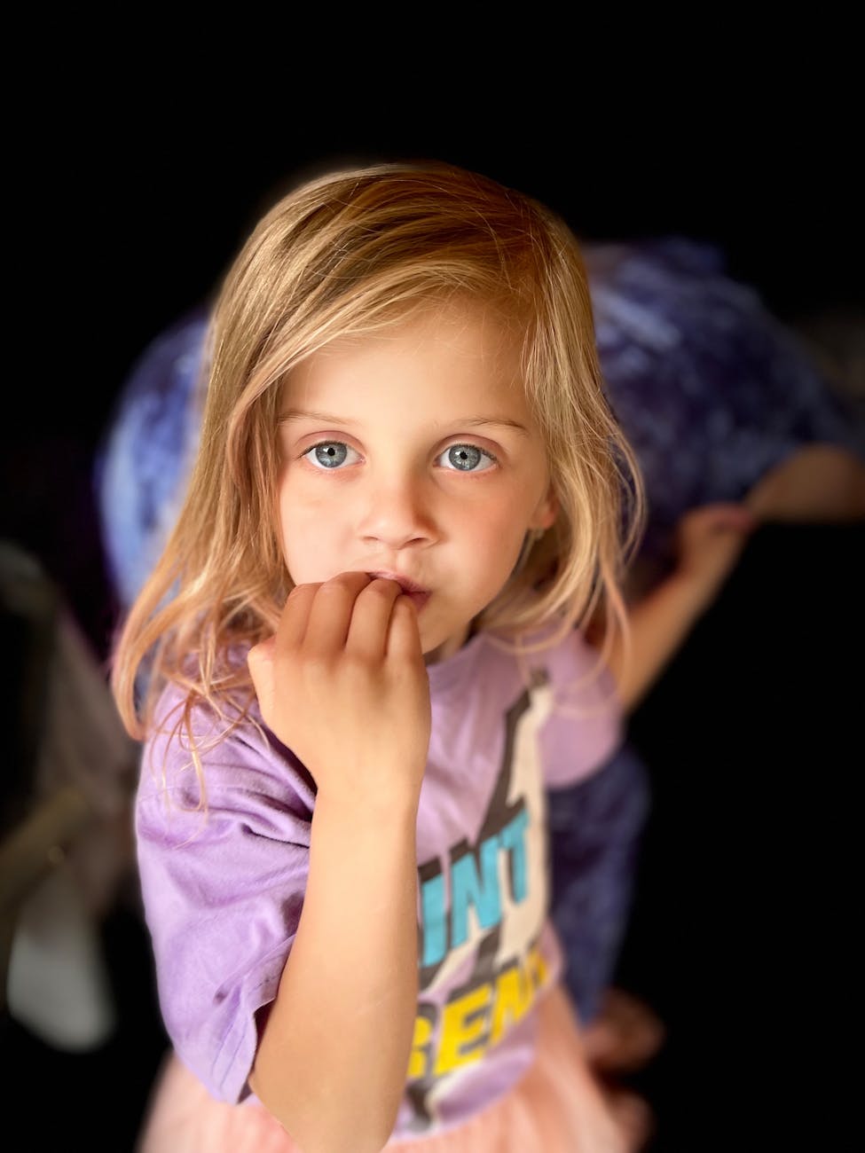 A young girl with long blonde hair is looking thoughtfully at the camera, wearing a purple t-shirt and staring with blue eyes while holding her hand to her mouth.
