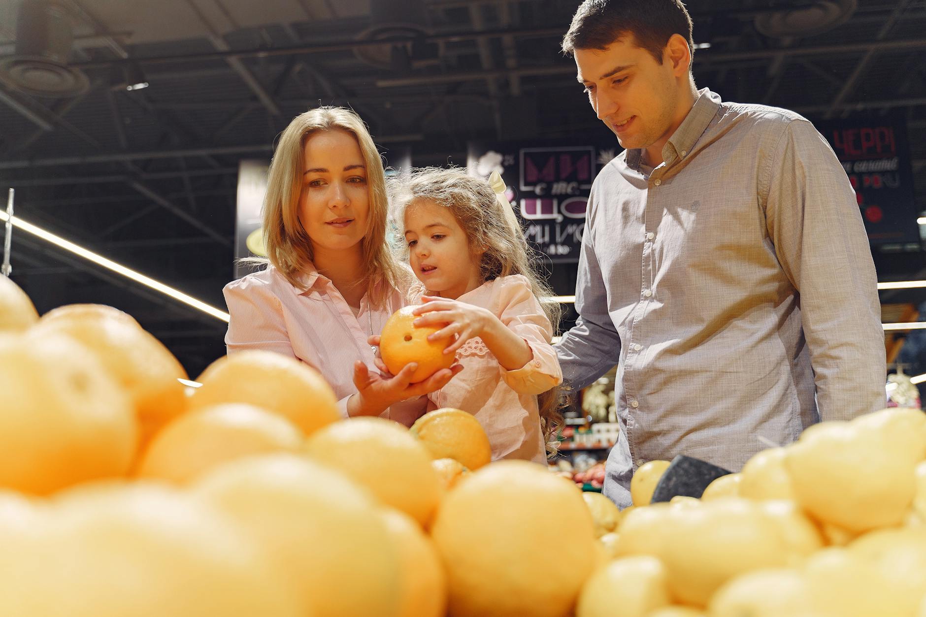 A family of three, including a woman, a girl, and a man, shopping for oranges at a market. The woman holds an orange while the girl examines it, with a display of oranges in the foreground.