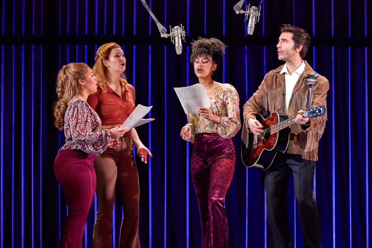 Four singers performing on stage with microphones, holding sheets of music, and accompanied by a guitarist.