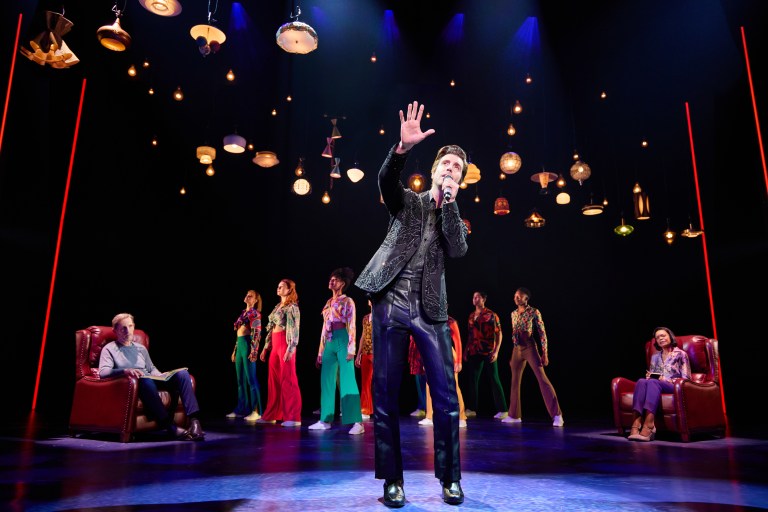 A performer in a sparkling jacket sings passionately on stage, surrounded by dancers wearing colorful outfits, with a vintage armchair and an audience member off to the side.