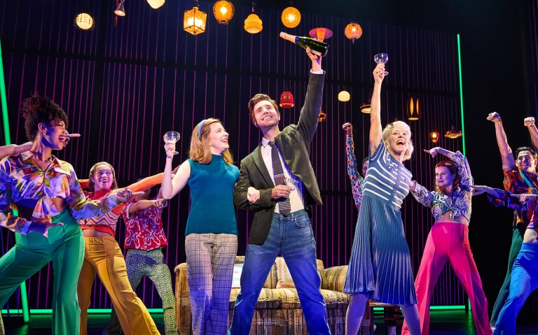 A lively musical scene featuring a group of performers celebrating with drinks, dressed in colorful outfits, in front of a backdrop with decorative lighting.