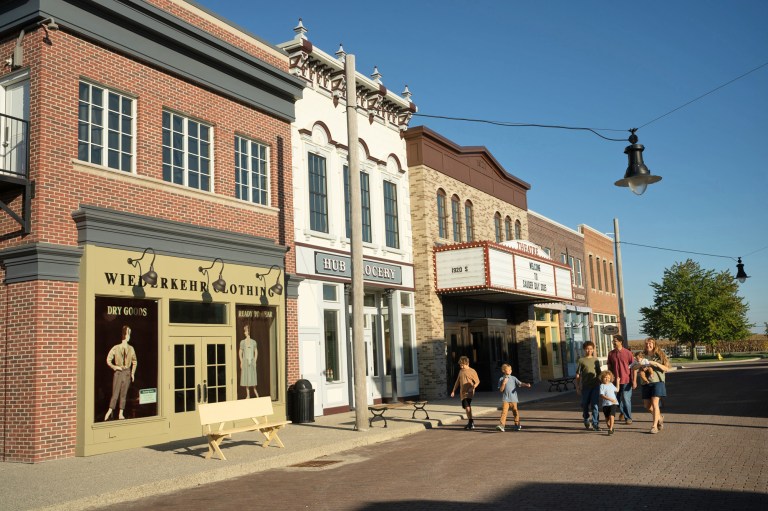A vibrant street scene featuring people walking in front of historic storefronts, including a clothing shop and a grocery store, under a clear blue sky.