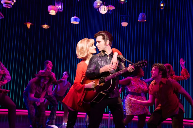 A couple embraces on stage, with the man playing an acoustic guitar and the woman wearing a red dress. The background features colorful lighting and a group of dancers performing energetically.