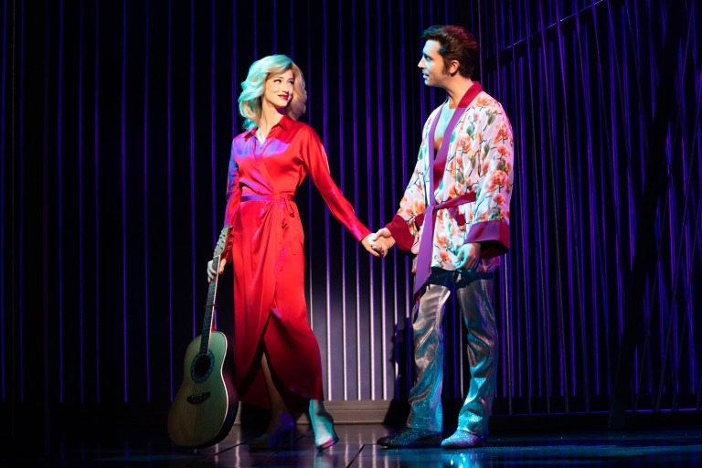 A scene from a musical featuring a woman in a red dress holding an acoustic guitar and a man in a colorful robe and shiny pants, both holding hands on stage.