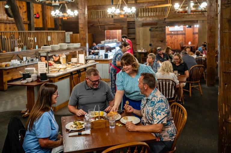 A lively restaurant scene featuring a group of four people dining at a wooden table, with a waitress serving food. The interior is rustic with wooden beams and warm lighting, and other patrons can be seen enjoying their meals in the background.