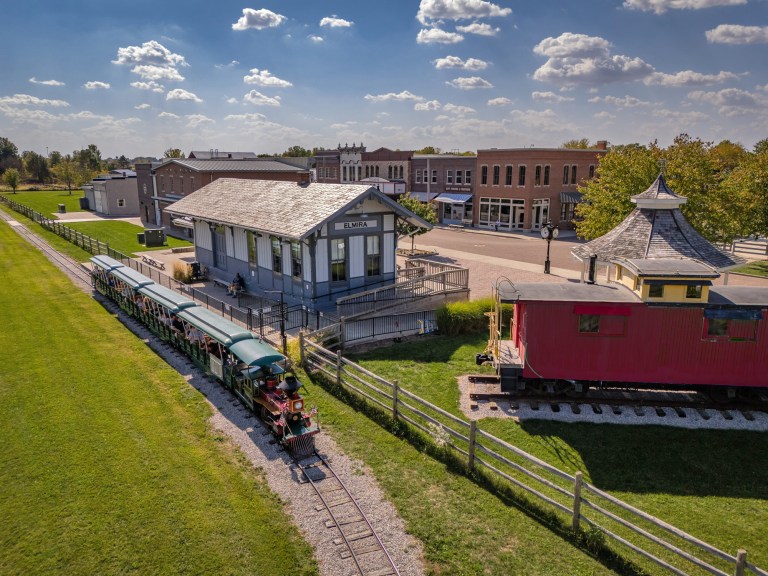A scenic view of a vintage train station labeled 'Elmira', with a train on the tracks and historic buildings in the background, under a partly cloudy sky.