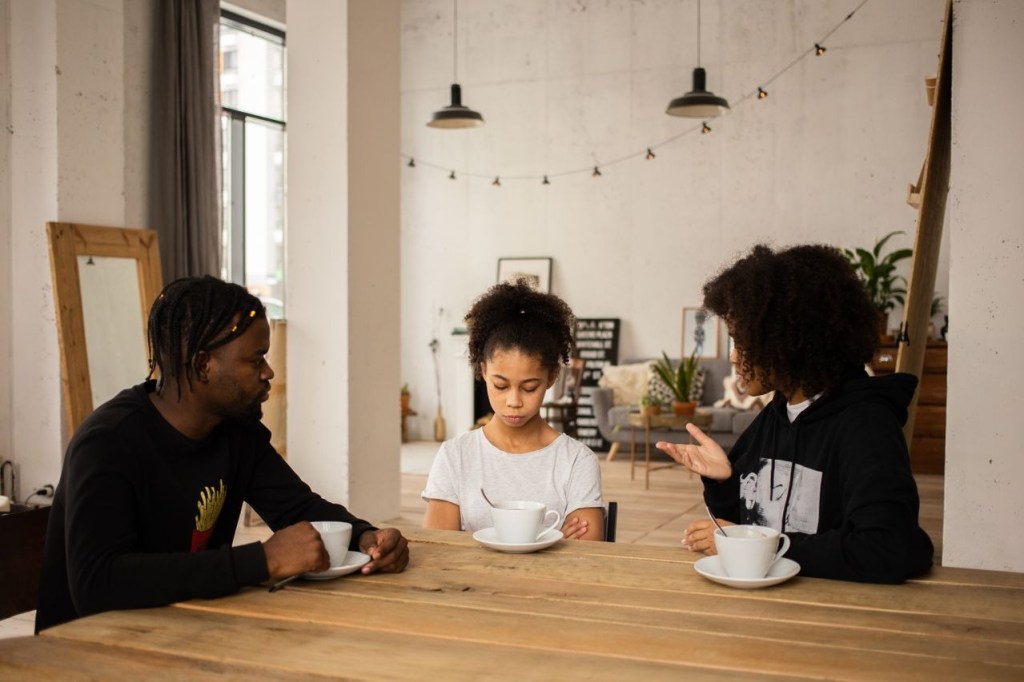 A man and a woman sit at a table with a young girl in a cozy, modern interior. The girl appears contemplative, while the woman gestures animatedly. Each person has a cup of coffee or tea.