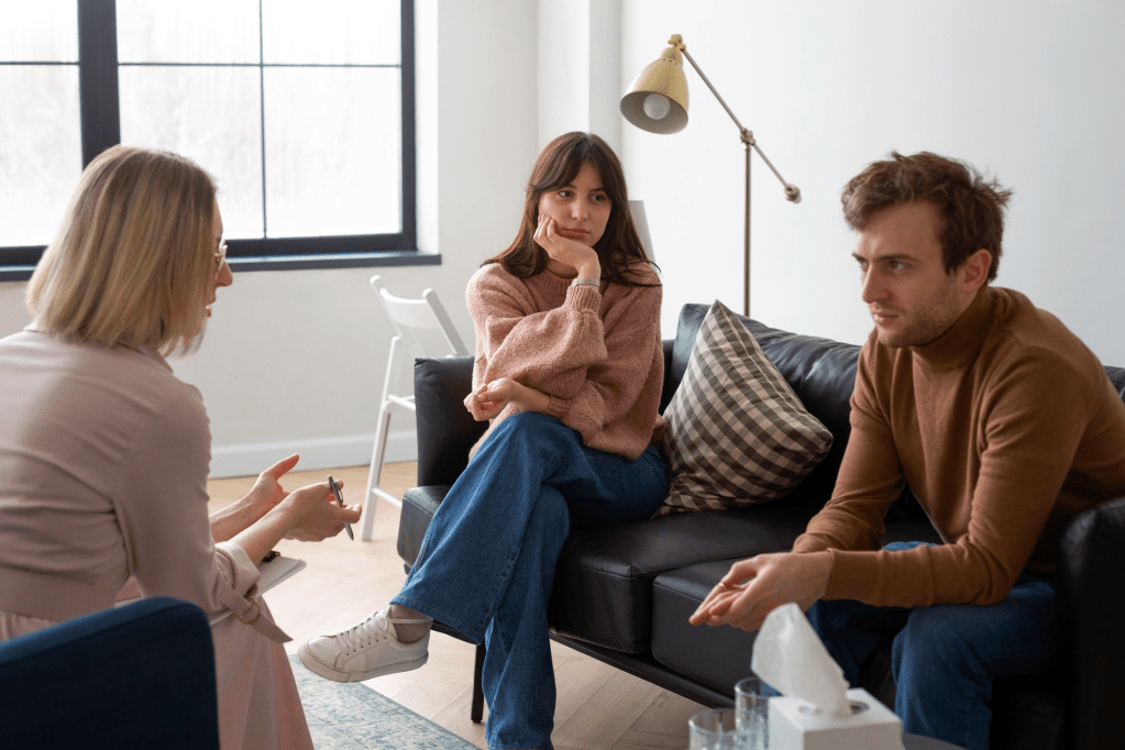 A therapy session featuring three individuals. Two people, one wearing a pink sweater and another in a brown turtleneck, are seated on a couch, looking contemplative. A third person, with blonde hair and glasses, is facing them, engaged in discussion.