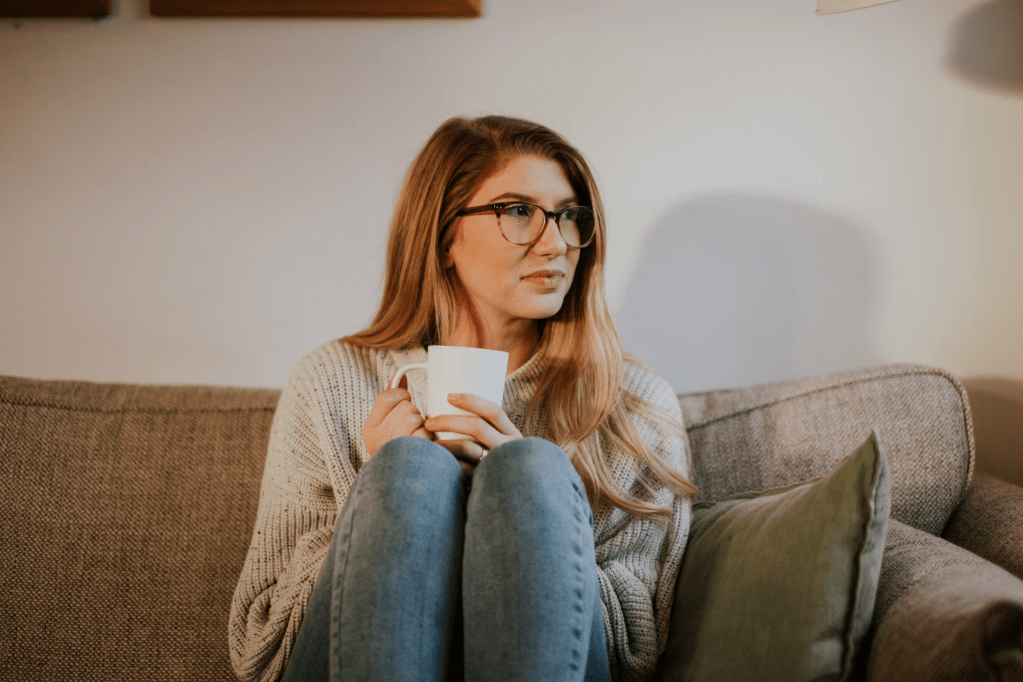 A woman with long hair and glasses sits on a couch, holding a cup of coffee, gazing thoughtfully into the distance.