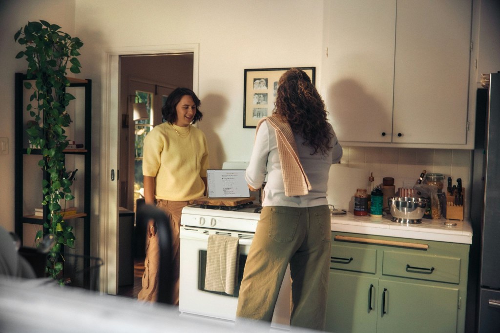 Two women in a cozy kitchen, one wearing a yellow sweater and the other in a gray top, sharing a joyful moment while cooking. A houseplant is visible in the background.