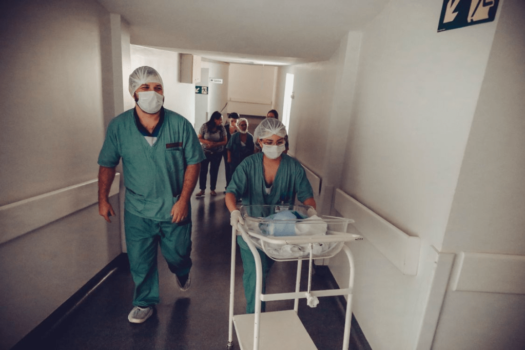 Two healthcare workers in scrubs and masks walking in a hospital hallway, one pushing a cart with medical supplies.