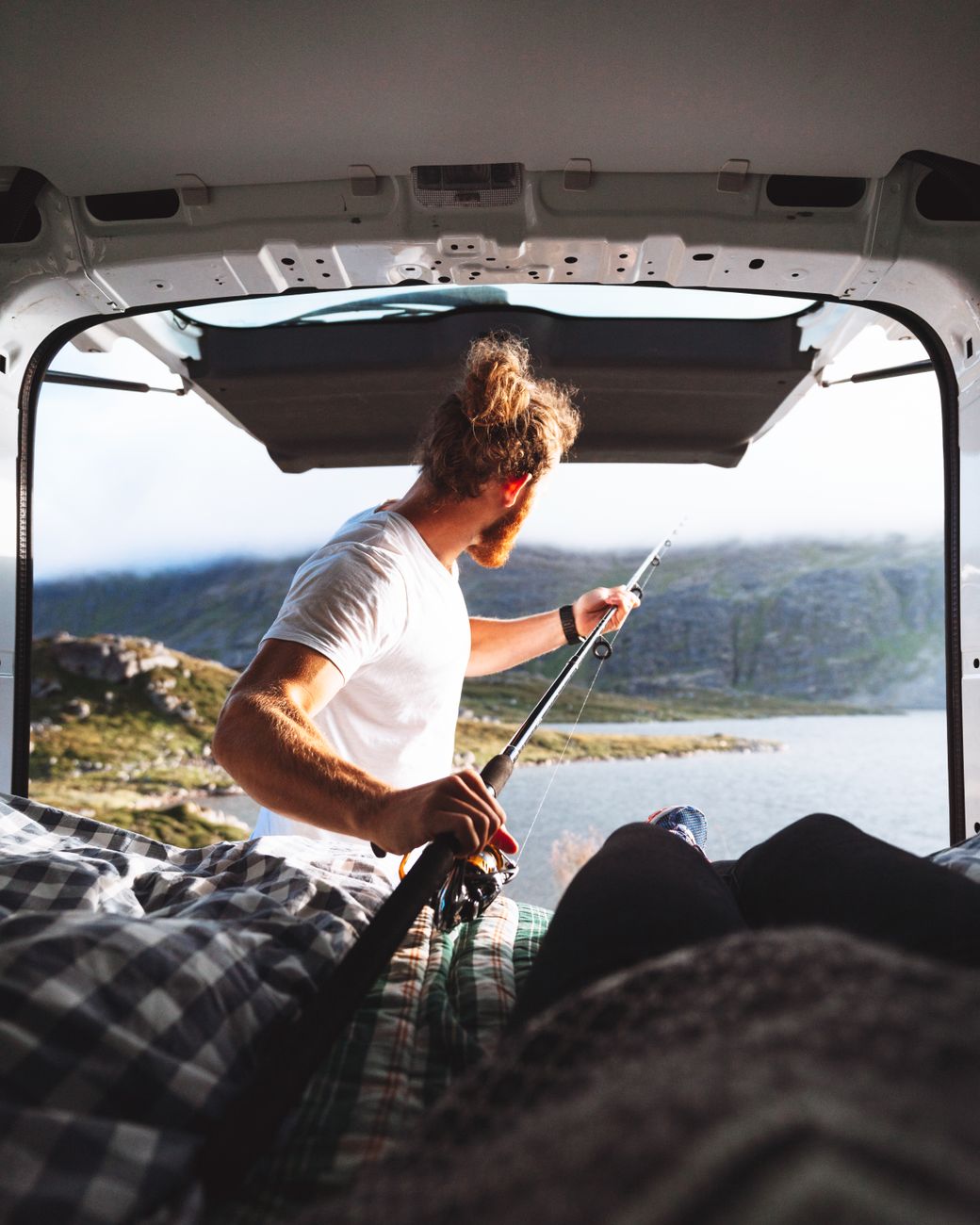 A person with long hair and a beard holding a fishing rod, looking out from the back of a van toward a scenic lakeside view surrounded by mountains.