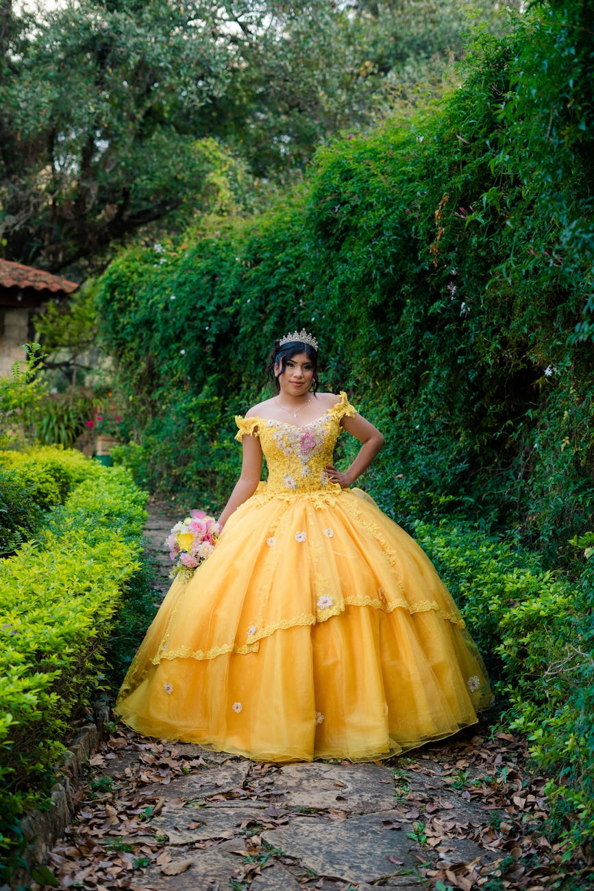 A young woman in a bright yellow gown with floral embellishments poses in a garden setting, surrounded by greenery and a stone pathway.