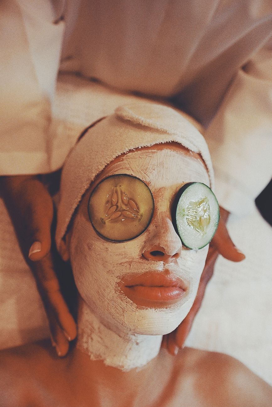 A person receiving a facial treatment, wearing a white facial mask and cucumber slices over their eyes, with hands gently applying the facial treatment.