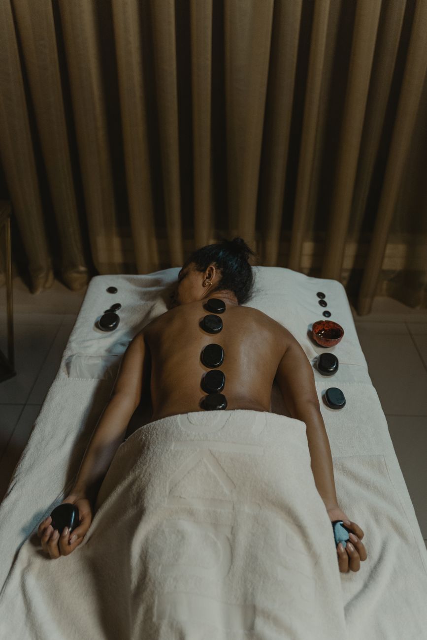 A person receiving a hot stone massage, laying on a towel with several smooth black stones placed along their back.