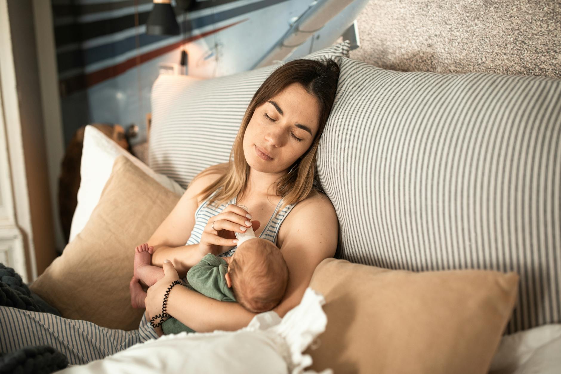 A woman sitting on a bed holds and gently feeds a baby with a bottle, surrounded by soft pillows and a cozy setting.