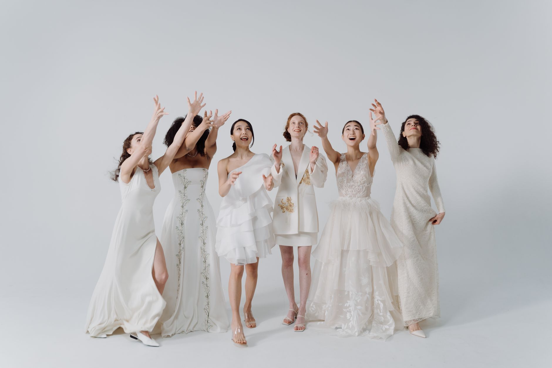 A group of seven women wearing a variety of white dresses, joyfully reaching upwards and smiling against a light gray background.