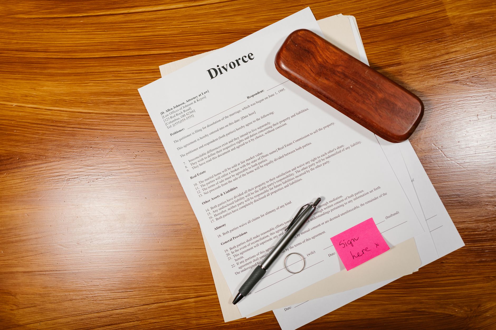 A divorce document placed on a wooden table, with a silver pen, a wooden case, and a sticky note that says 'Sign here'.
