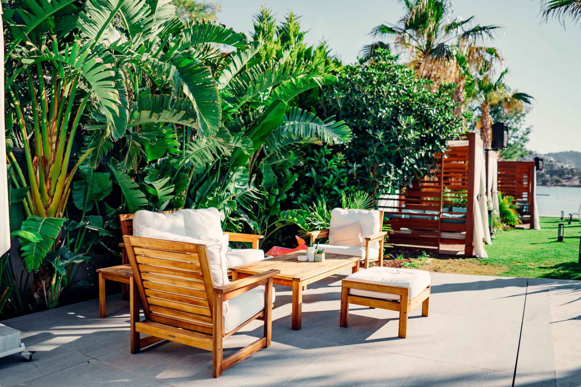 A cozy outdoor seating area featuring wooden furniture with white cushions surrounded by tropical greenery.