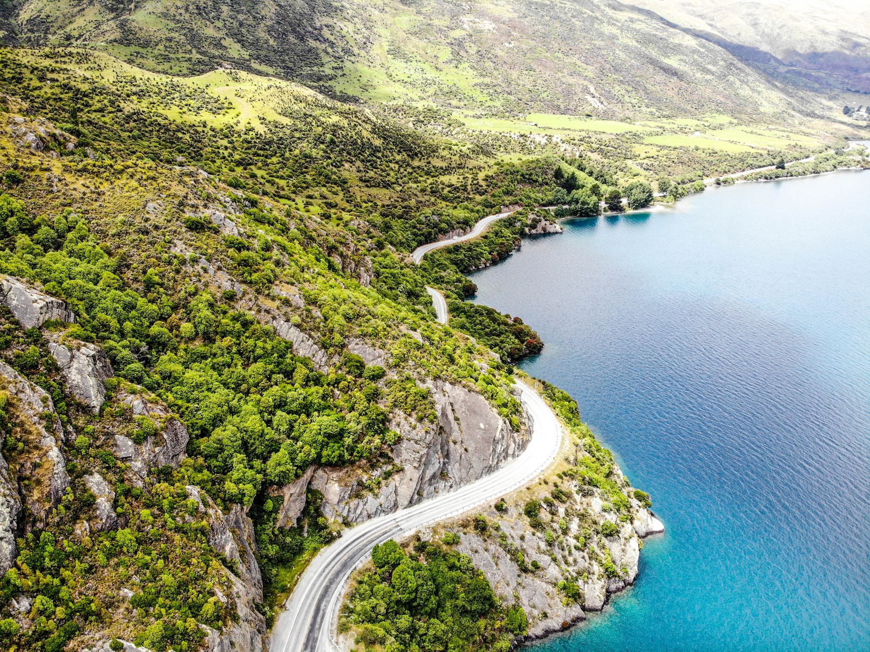 Aerial view of a winding road along a vibrant green mountainous landscape beside a blue lake.