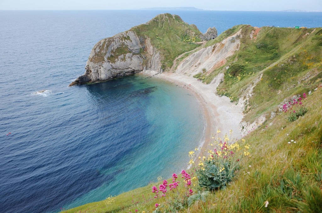A scenic coastal view featuring a crescent-shaped beach, clear blue waters, and grassy cliffs adorned with wildflowers.