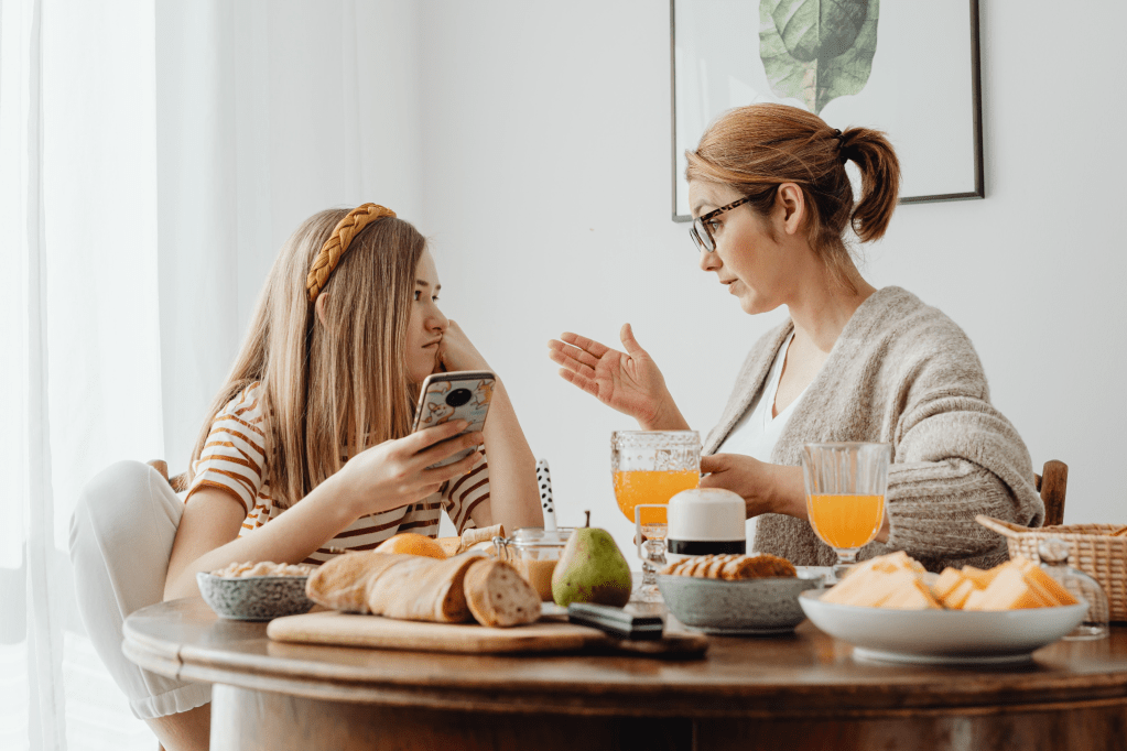 A mother and daughter engaged in conversation over breakfast at a wooden table filled with food and drinks, with the daughter looking at her phone.