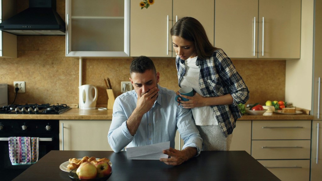 A man sitting at a table in a kitchen reads a letter with a concerned expression while a woman stands beside him, holding a cup and looking at the letter.