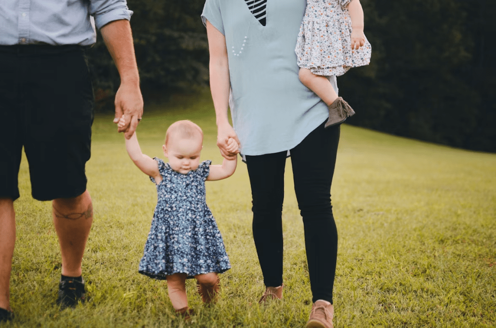 A family standing on grass, holding hands. One adult is wearing a blue shirt and black shorts, while the other adult is wearing a light top with a slit and black pants. A young child in a blue floral dress stands between them, holding their hands.