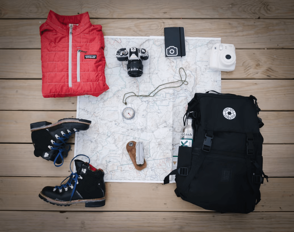 Flat lay of outdoor gear including a red insulated jacket, black hiking boots, a map, a compass, a camera, a smartphone, a small white camera, a multitool, and a black backpack with a water bottle.