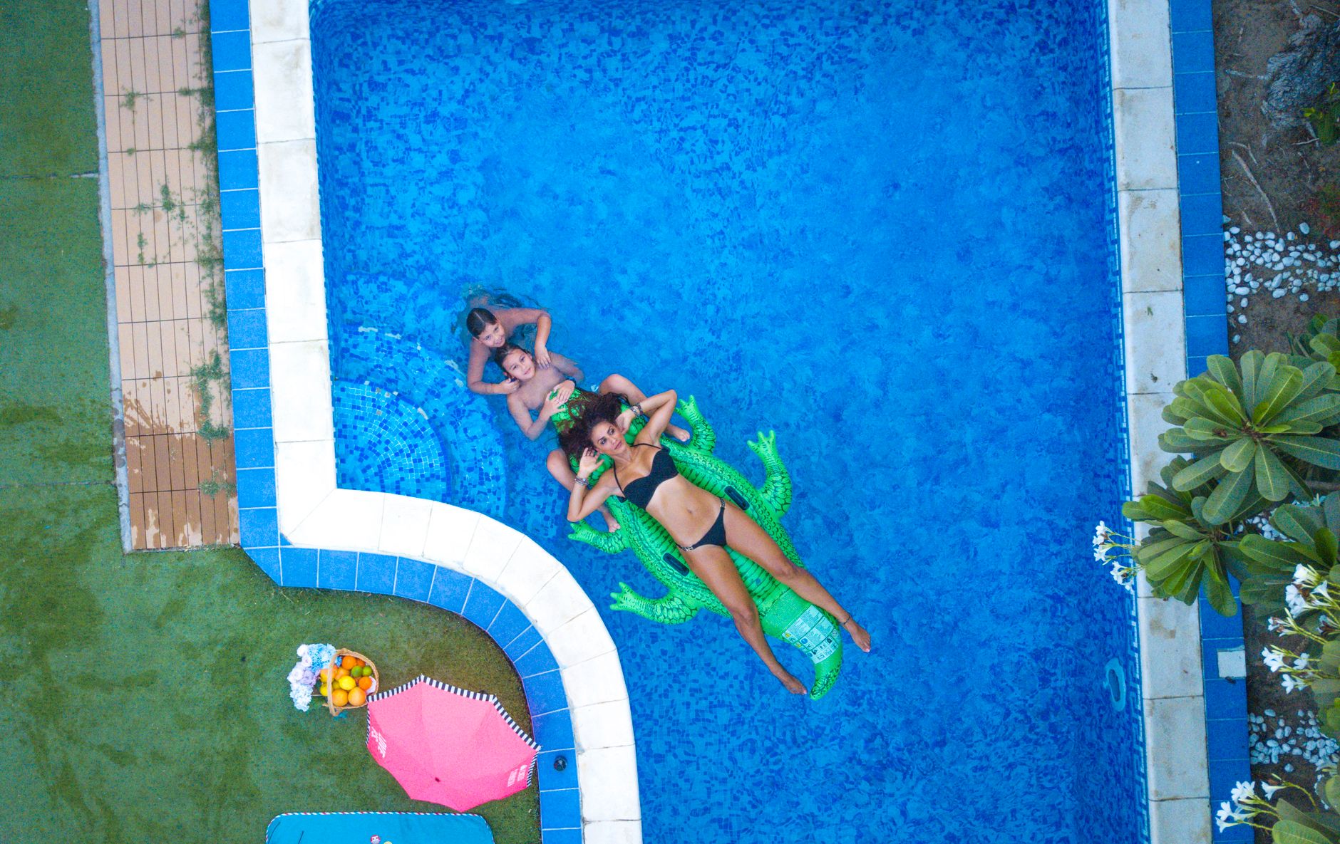 Aerial view of three people in a blue swimming pool, one lounging on a green alligator float while two others are seated nearby. Surrounding the pool are green grass and a pink beach umbrella.
