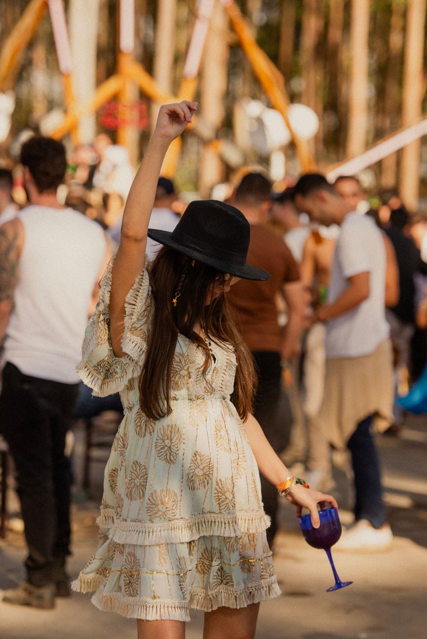 A woman in a light floral dress and a black hat dances with one hand raised, holding a blue drink, surrounded by a crowd at an outdoor event.