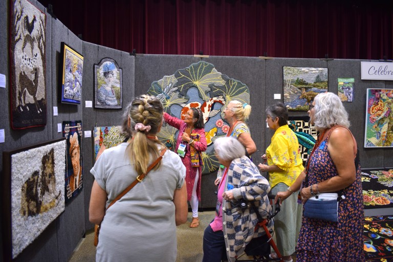 A group of women admiring and discussing textile art at an exhibition, featuring various colorful artworks hanging on the wall.