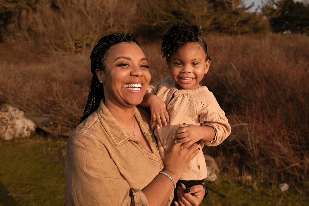 A smiling woman holding a young girl outdoors, surrounded by grass and trees, both enjoying a sunny day.