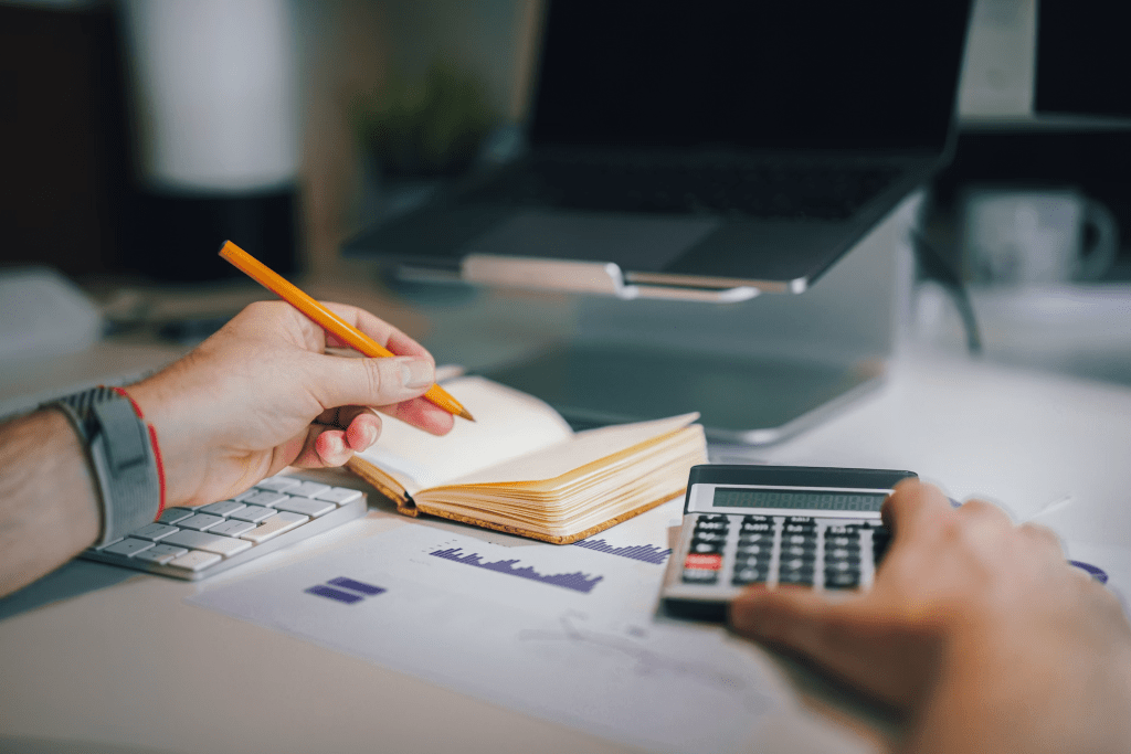 A person using a pencil to write in a notebook while a calculator and a keyboard are visible on a desk.