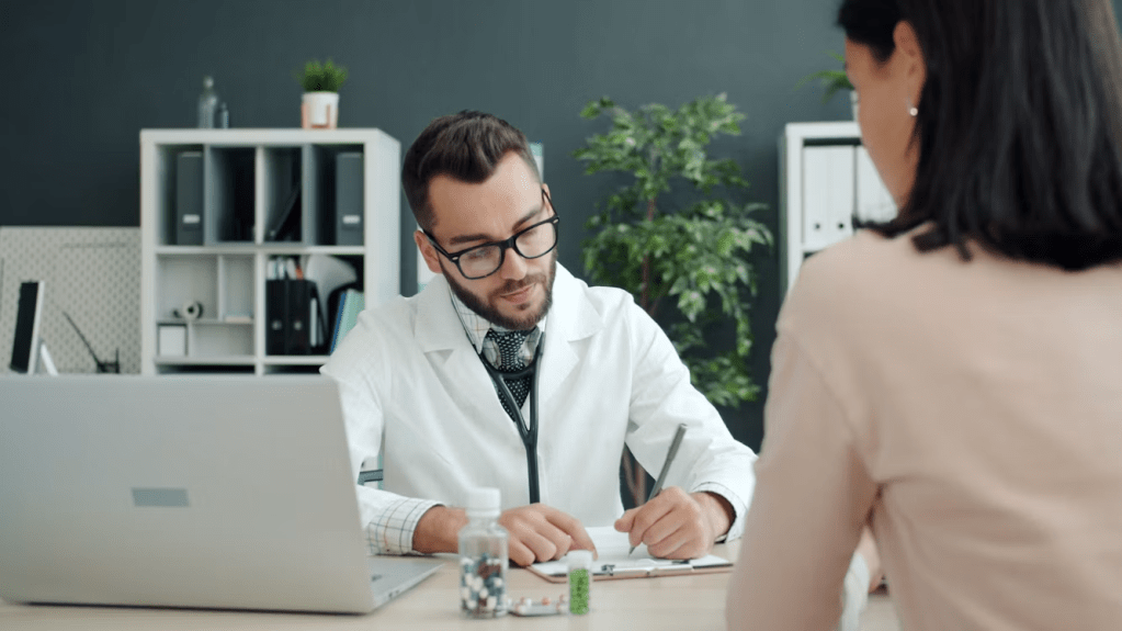 A male doctor in a white coat is sitting at a desk, taking notes while consulting with a female patient who is facing him.
