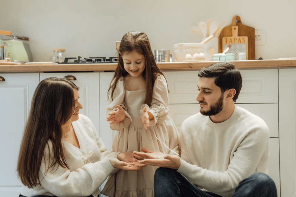 A family of three, including a mother, father, and young girl, playing together in a kitchen. The girl is smiling and clapping her hands while the parents look on affectionately.