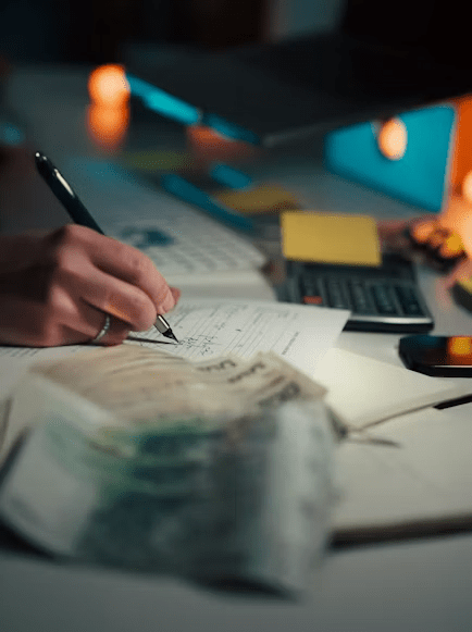 A person writing notes with a pen on a notepad, surrounded by a calculator, a mobile phone, and a stack of money on a desk, with soft lighting in the background.