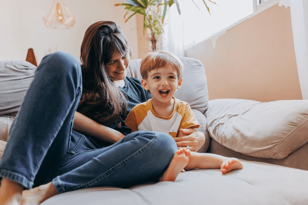 A mother sitting on a couch with her young son, both smiling and enjoying a moment together in a cozy living room.