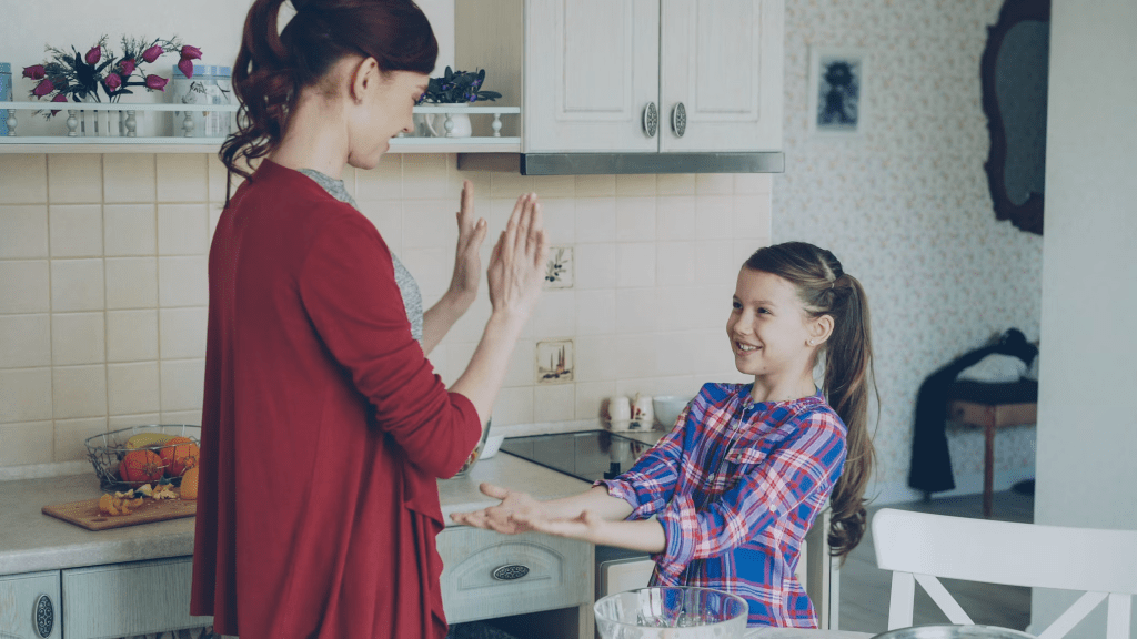 A mother and daughter playfully interacting in a kitchen, with the mother clapping her hands and the girl smiling while reaching out her hands.
