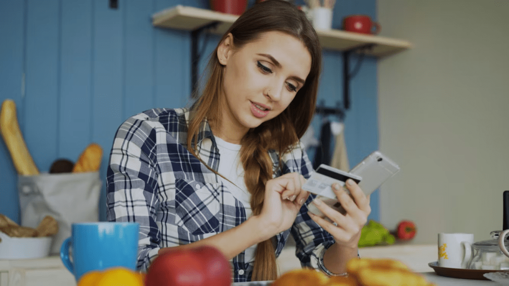 A young woman in a plaid shirt sitting at a kitchen table, looking at her smartphone while holding a credit card, with breakfast items and a blue mug in the foreground.
