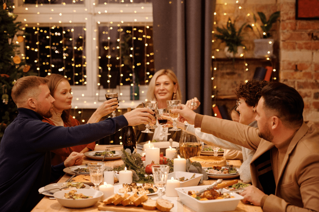 A group of six people joyfully toasting with drinks around a festive dinner table adorned with candles, plates of food, and Christmas lights in the background.
