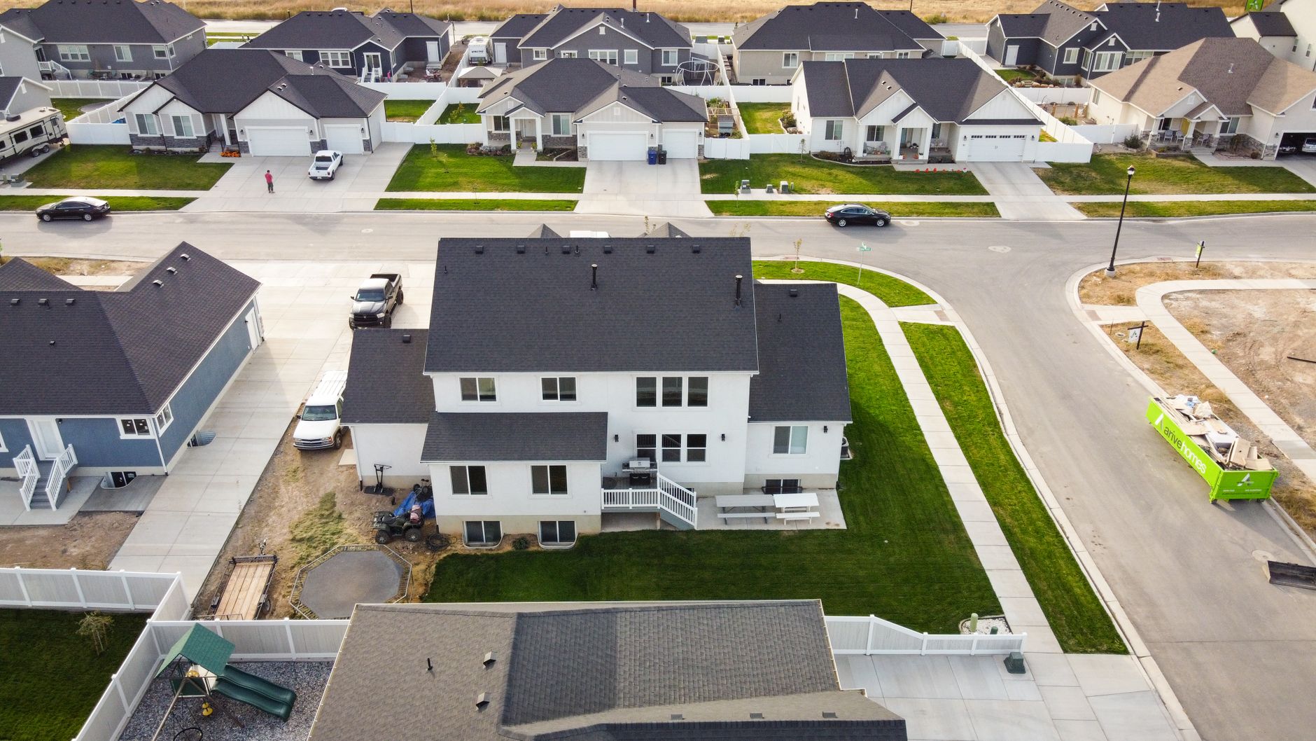 Aerial view of a suburban neighborhood featuring several houses of varying designs, with a focus on a large white house with a black roof, a neatly maintained lawn, and a patio area. Nearby, several parked cars are visible, along with a playground slide in a fenced yard.