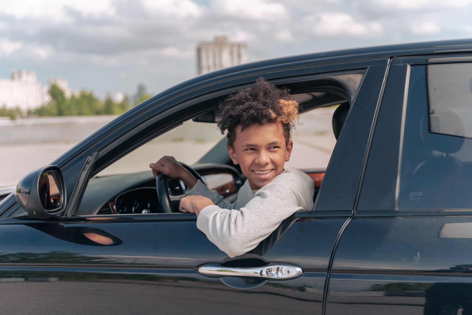 A young boy with curly hair pokes his head out of the window of a black car, smiling while resting his arm on the car door.
