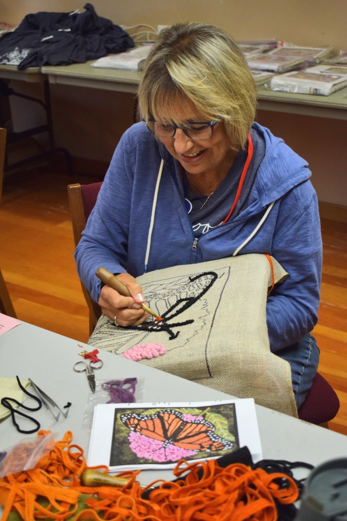 A woman with short blonde hair working on a textile craft, smiling as she uses a punch needle to create a butterfly design on burlap fabric. Scissors and colorful threads are visible on the table.