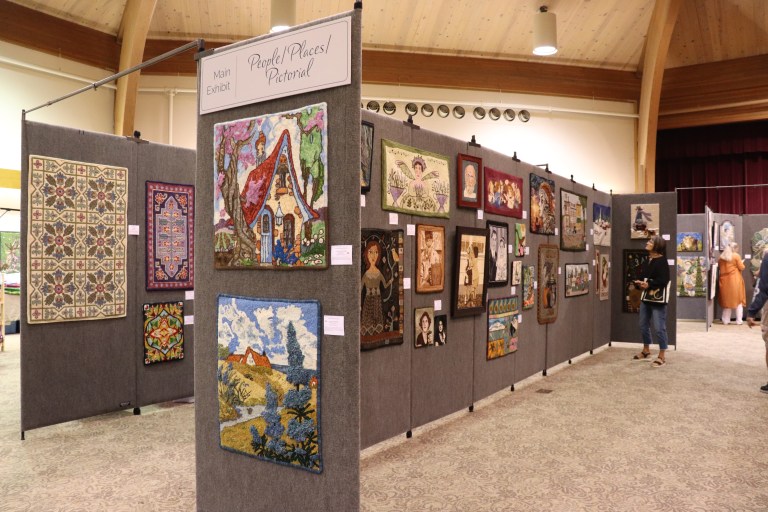 An art exhibit featuring various textile artworks, including rugs and tapestries, displayed on partition walls in a spacious hall.