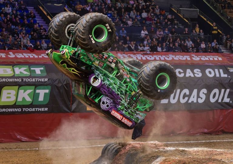 A monster truck performing a wheelie in an arena, with a crowd in the background and dust flying from the ground.