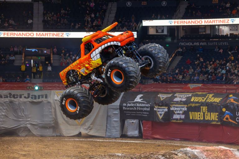 A brightly painted monster truck performing a jump in an arena during a motorsport event, with an excited crowd in the background.