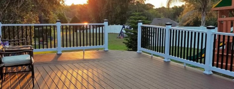 A spacious wooden deck with black and white railing overlooking a green backyard at sunset, featuring chairs and playground equipment in the distance.
