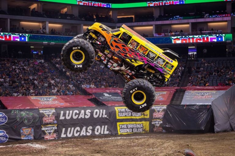 A monster truck performing a jump in a stadium during a live event, with a crowd in the background and vibrant advertisements visible.